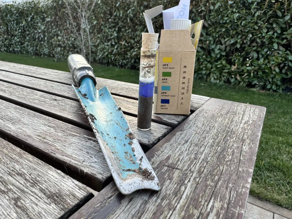 Mein Setup zum pH-Bodentest: Der Gardena Unkrautstecher, der geöffnete pH-Neudorff-Bodentest liegen auf meinem Gartentisch. Davor sieht man das Ergebnis des pH-Bodentests im Röhrchen - mit einer dunkelblauen Färbung.