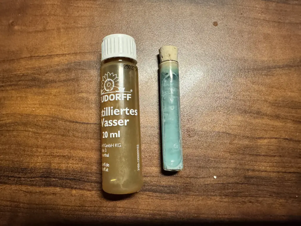 Nahaufnahme des Neudorff pH-Bodentest-Sets: Ein Röhrchen gefüllt mit der cyanfarbenen Kontroll-Flüssigkeit nach Zugabe von destilliertem Wasser und Pulver, daneben die Originalflasche des destillierten Wassers auf Holzuntergrund. 