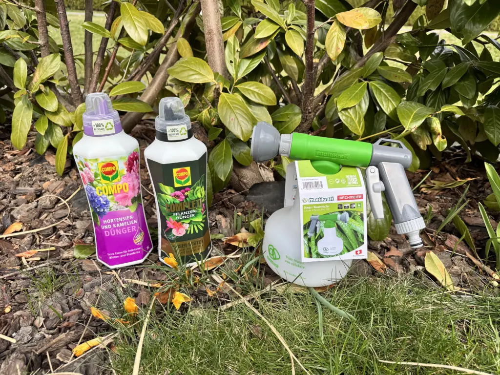 Zwei Flaschen Flüssig-Dünger mit einem Birchmeier Aquamix stehen auf dem Boden im Garten vor einer Kirschlorbeerhecke