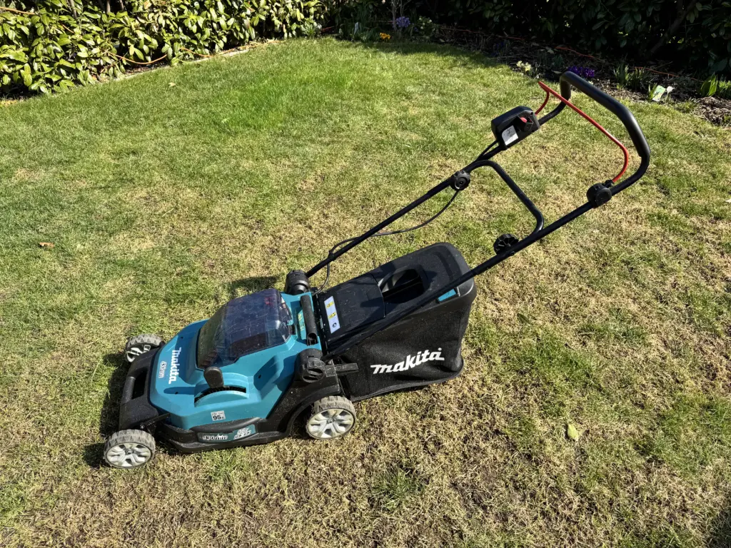 Der Akku-Rasenmäher Makita DLM432 steht auf meinem Rasen - im Hintergrund ist etwas Hecke und ein Beet zu sehen.