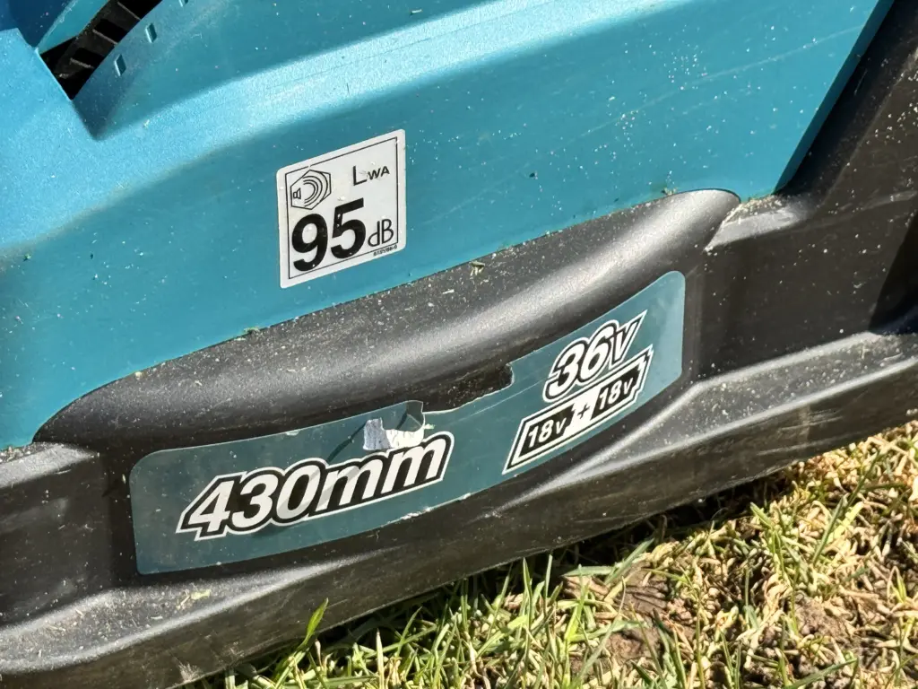 Die linke Seite des Makita DLM432 Akku-Rasenmähers ist etwas beschädigt. Auf den Aufklebern des Rasenmähers kann man "LWA 95dB" und "430 mm" und "18V+18V=36V" lesen