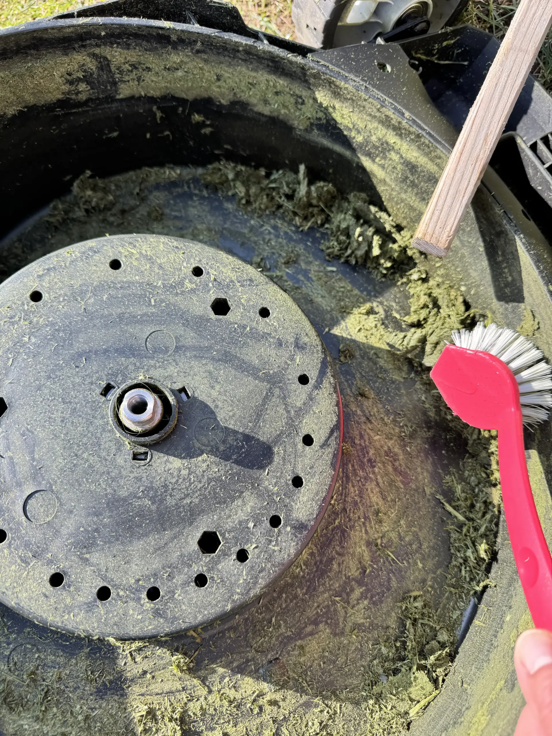 Das Mähdeck des Makita DLM432 - immer noch schmutzig, aber die ersten Grasreste vom letzten jahr liegen lose im unteren Teil - auf dem Foto sind eine Bürste und ein Holz zu sehen, mit dem die Grasreste abgekratzt werden.
