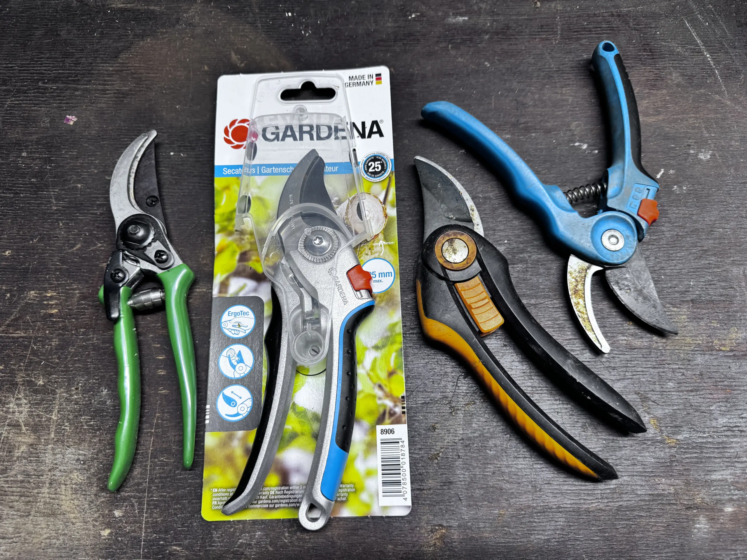 Verschiedene Gartenscheren im Test auf einer Werkbank: Eine grüne No-Name Schere, eine neue Gardena Bypass Schere 8906 und zwei Modelle von Fiskars