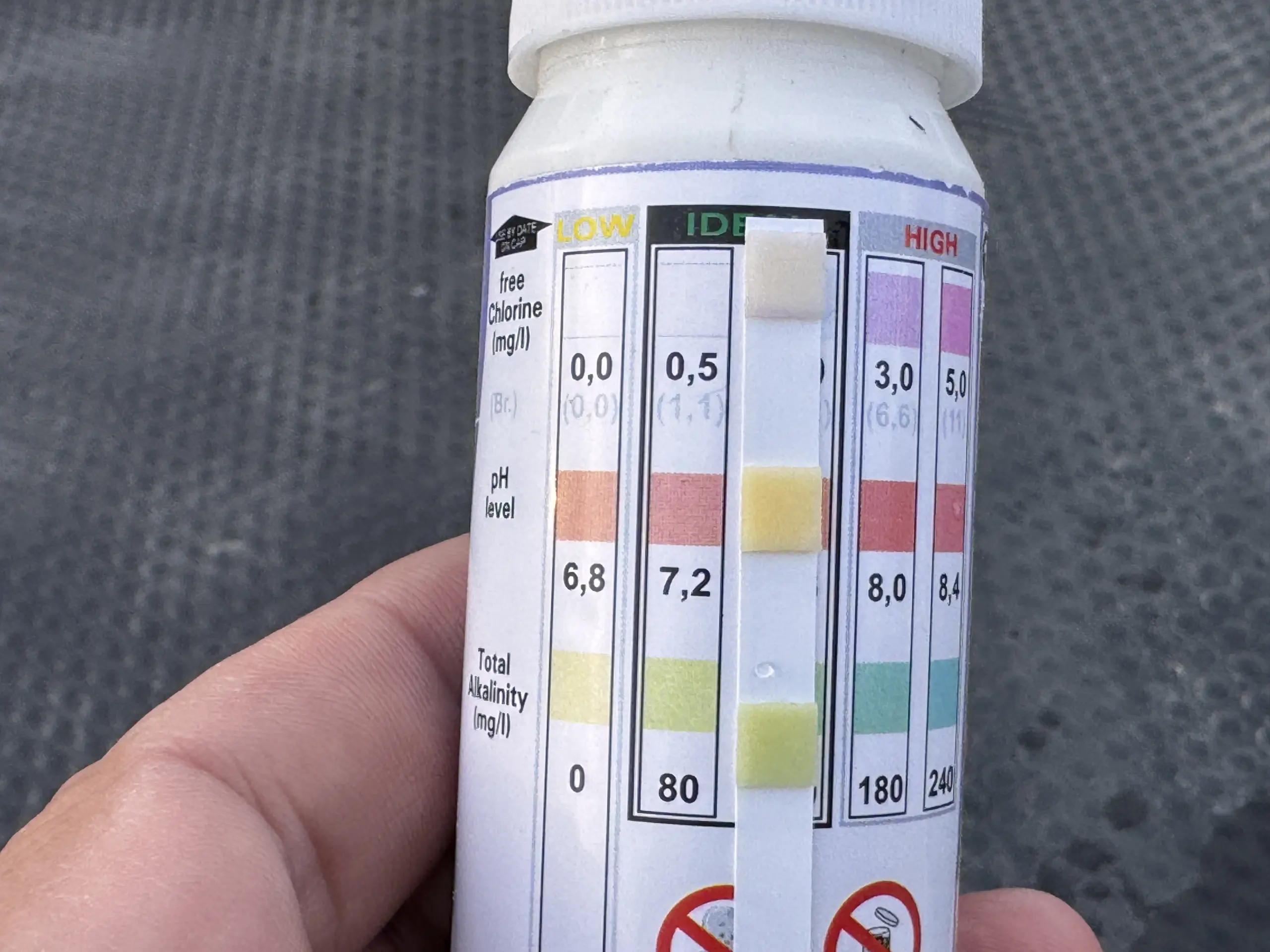 Poolwasser einstellen mit einem Stäbchentest - das Stäbchen zeigt ein Ergebnis vor dem Hintergrund der Skala auf dem Behälter an - man kann kaum erkennen, welcher pH-Wert abzulesen ist.an