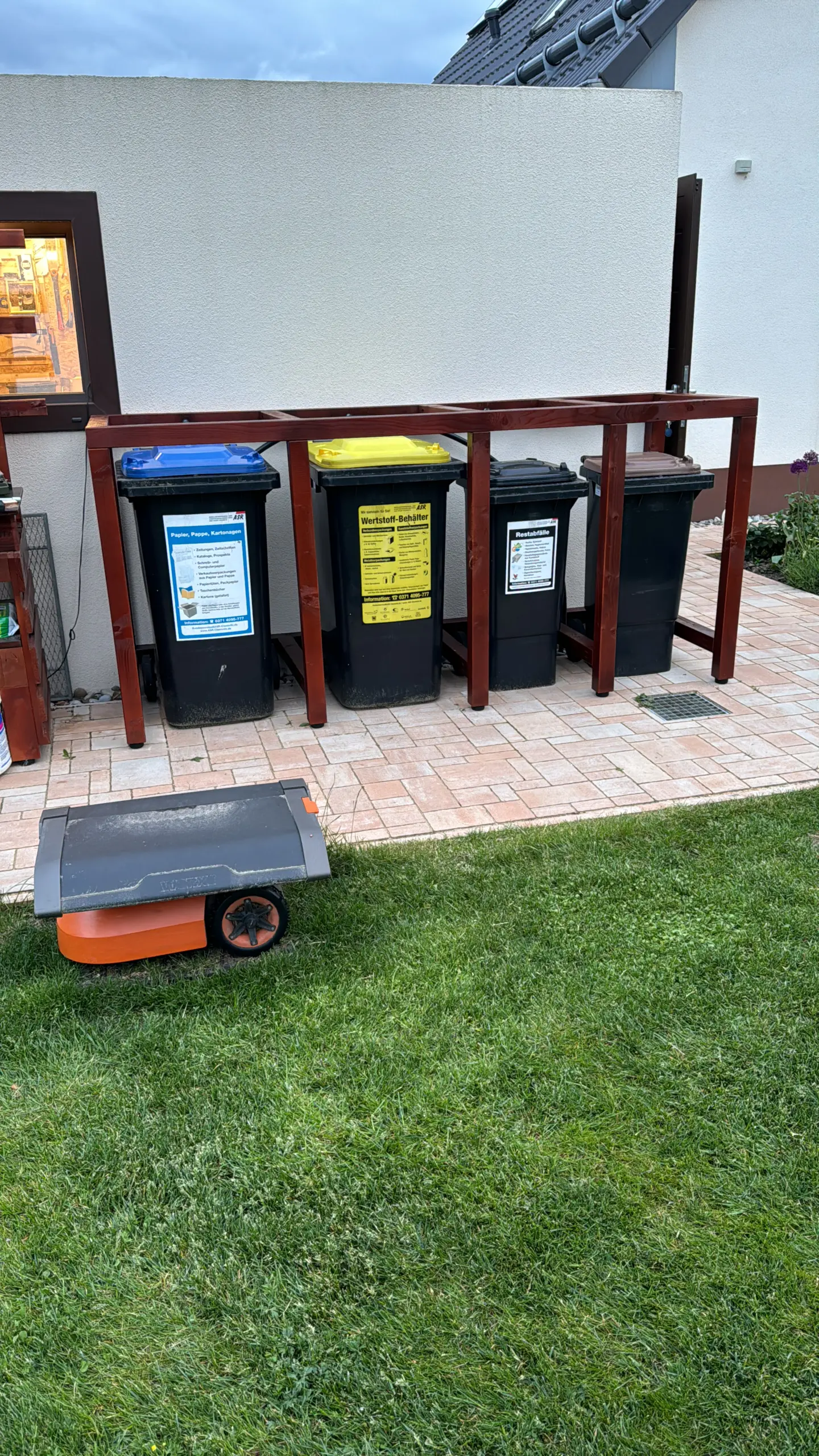 Ein Mähroboter mit Begrenzungskabel steht in der Garage vor einem Schuppen mit einer Konstruktion für die Mülltonnenbox