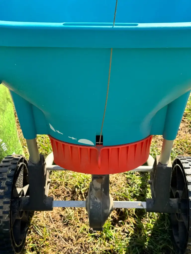 Unterseite meines blauen Gardena XL Schleuderstreuers mit rotem Stellrad, weißer Richtungsmarkierung und Seilzug zwischen den Laufrädern