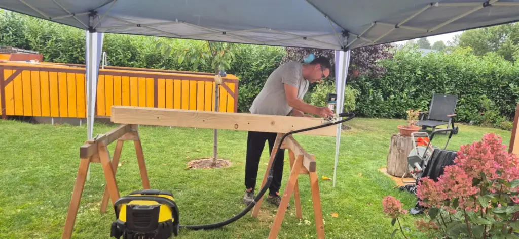 Handwerker beim Fräsen eines Holzbalkens mit der Makita DRT50 und angeschlossenem Absaugschlauch auf Böcken im Garten