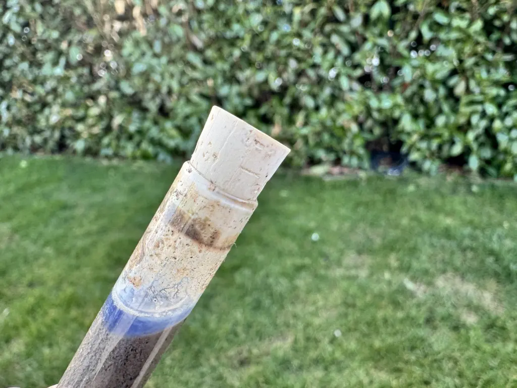 pH-Bodentest Ergebnis: Nahaufnahme eines Teströhrchens mit blauer Lösung und abgesetzter Erde vor grünem Gartenhintergrund