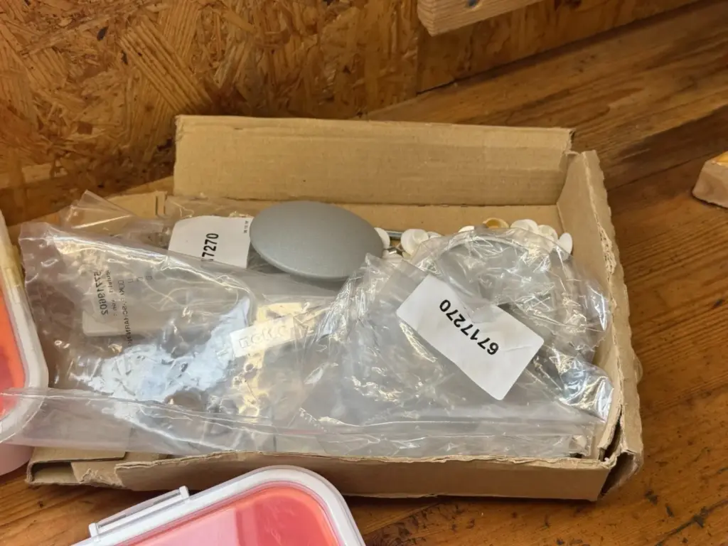 Offener Karton mit Beschlägen, Kunststoffteilen und beschrifteten Beuteln auf einer Holzablage.