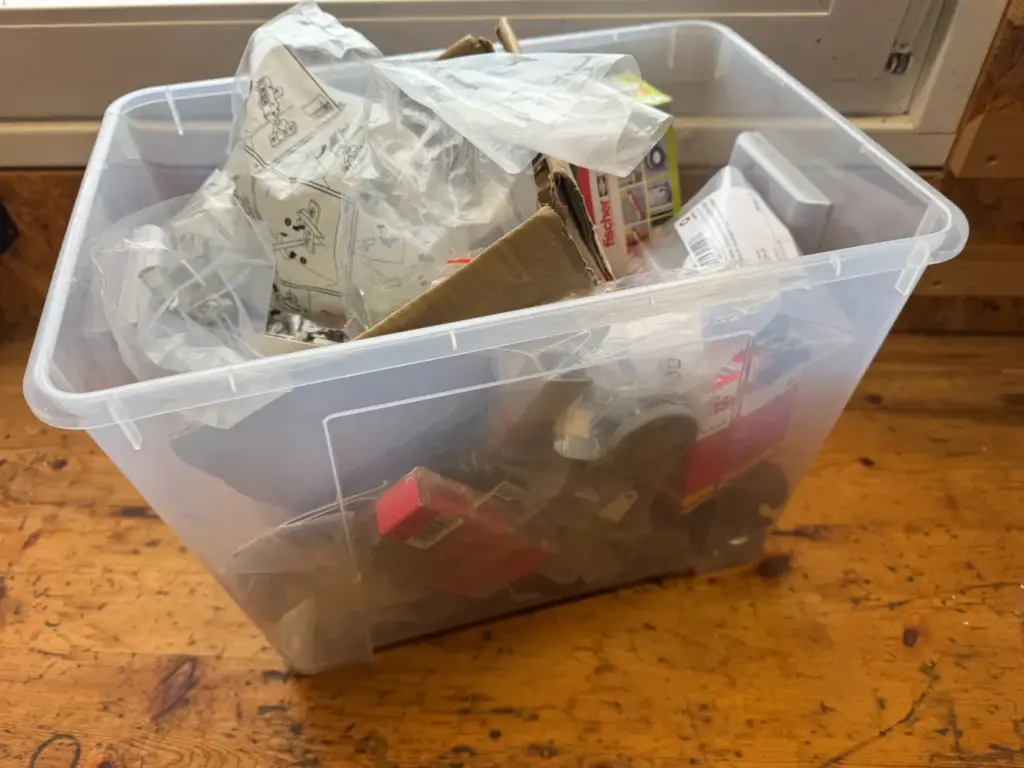 Schräger Blick auf eine offene transparente Sammelbox mit Kartons, Beuteln und Beschlägen.